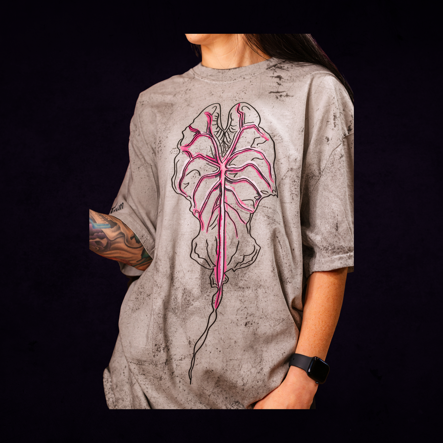 Person wearing a gray t-shirt with a pink and black Alocasia Venom leaf design on a dark background